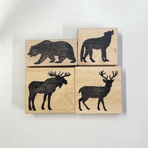 Puede incluir: Cuatro bloques de madera con imágenes en silueta negra de un oso, un lobo, un alce y un ciervo. Los bloques son todos del mismo tamaño y forma.