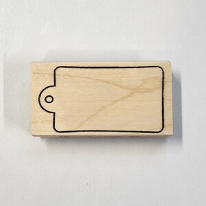 May include: A wooden stamp with a rectangular outline and a small circle for a hanging loop. The stamp is blank and ready to be used for crafting or scrapbooking.