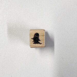 Op de afbeelding: Een houten stempel met een zwart spookontwerp. Het spook heeft twee ogen en een kleine mond.