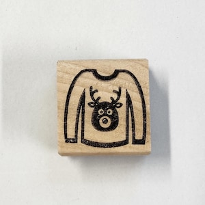 May include: A wooden stamp with a black and white design of a Christmas sweater with a reindeer on the front.