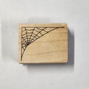 May include: A wooden stamp with a black spiderweb design in the corner. The stamp is made of light wood and has a rectangular shape.