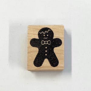 May include: A wooden stamp with a black gingerbread person design. The gingerbread person has a bow tie and a wavy line for hair.