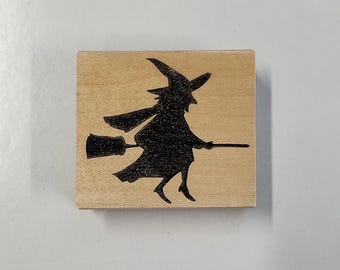 Könnte beinhalten: Ein Holzstempel mit einer schwarzen Silhouette einer Hexe, die auf einem Besen fliegt.