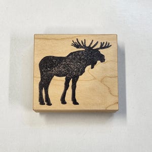 May include: A wooden block stamp featuring a black silhouette of a moose with antlers.