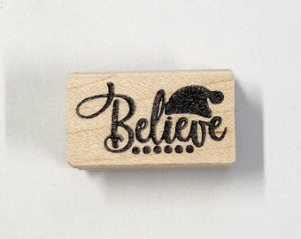 以下が含まれることがあります： 「Believe」という単語が筆記体で書かれた木製のスタンプ。単語の上にサンタ帽子のグラフィックが描かれている。