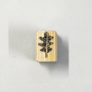 May include: A small, rectangular wooden stamp with a black oak leaf design. The stamp is made of light-coloured wood and is likely used for crafting or scrapbooking. The leaf is centered on the stamp.