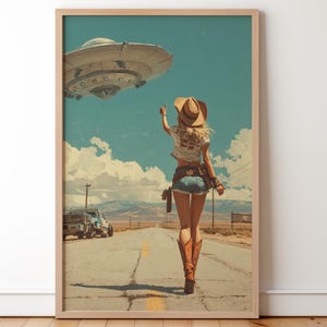 May include: Framed art print featuring a woman in a cowboy hat and boots walking down a desert road, gesturing towards a hovering UFO. The artwork has a vintage, retro style with a blue sky and a classic car.