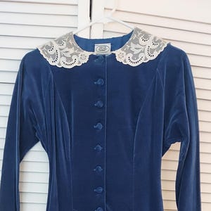 May include: A long, navy blue velvet dress with long sleeves and a button-down front. The dress features a decorative lace collar and a flared skirt, creating an elegant silhouette. The dress is hanging on a white hanger.