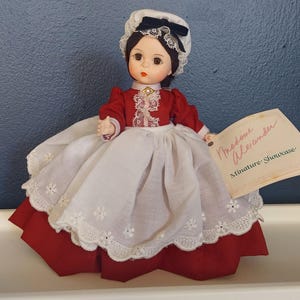 Puede incluir: Una muñeca de porcelana con un gorro blanco, un vestido rojo y un delantal blanco. La muñeca está etiquetada como "Madame Alexander Miniature Showcase".