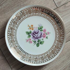 May include: White ceramic plate with a gold floral and geometric border. The center features a purple rose surrounded by pink and yellow flowers and green leaves. The plate has a gold rim.