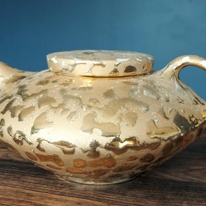 May include: A gold-colored teapot with a matching lid. The teapot has a rounded body, a curved handle, and a spout. The surface has a textured, mottled pattern. The teapot is sitting on a wooden surface.