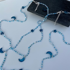 May include: A layered necklace with blue and white beads, featuring crescent moon and star charms. The necklace is made of small, delicate beads and has a simple, elegant design.