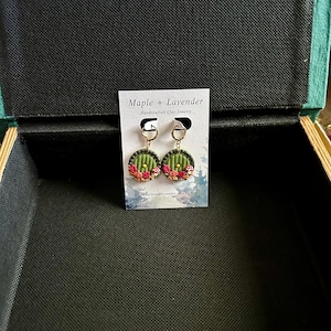 May include: A pair of green and red handmade clay earrings with a round shape. The earrings feature a small, detailed design of a door with a golden handle. The earrings are displayed on a white card with the text "Maple + Lavender Handcrafted Clay Jewelry".