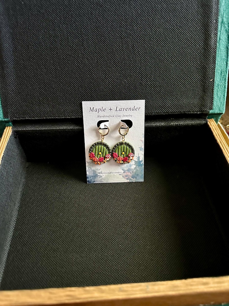 May include: A pair of green and red handmade clay earrings with a round shape. The earrings feature a small, detailed design of a door with a golden handle. The earrings are displayed on a white card with the text "Maple + Lavender Handcrafted Clay Jewelry".