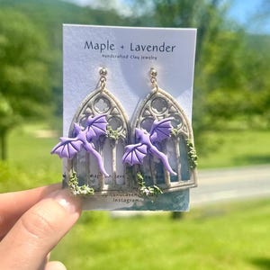May include: Handmade clay earrings with a gothic window design and a purple dragon. The earrings have green foliage and are held by a hand. The card reads "Maple + Lavender".