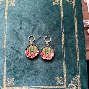 May include: A pair of green and gold earrings featuring a round, green door design. The door has a small, yellow circle in the centre and is surrounded by a brown frame. The earrings are decorated with small, red and orange mushrooms and leaves.