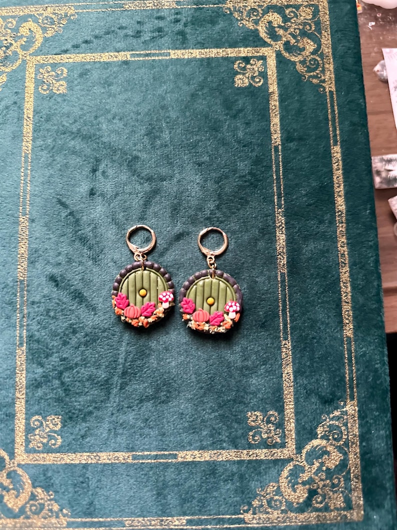 May include: A pair of green and gold earrings featuring a round, green door design. The door has a small, yellow circle in the centre and is surrounded by a brown frame. The earrings are decorated with small, red and orange mushrooms and leaves.