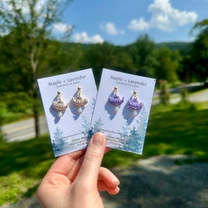 Może przedstawiać: Dwie pary ręcznie robionych kolczyków z gliny są prezentowane na tle błękitnego nieba i drzew. Jedna para ma złoty wzór czajniczka z perłową kroplą, a druga fioletowy czajniczek z podobną perłą. Na kartach widnieje napis "Maple + Lavender."