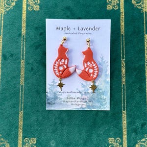 May include: A pair of red fox-shaped earrings with white floral details and gold star charms. The earrings are handmade from clay and are displayed on a white card with the text "Maple + Lavender Handcrafted Clay Jewelry" and "mapleandlavender.etsy.com Follow along at Mapleandlavender on Instagram."