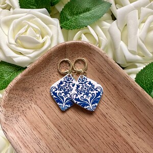 May include: A pair of blue and white patterned earrings with gold hoops. The earrings are shaped like a diamond and feature a floral design.