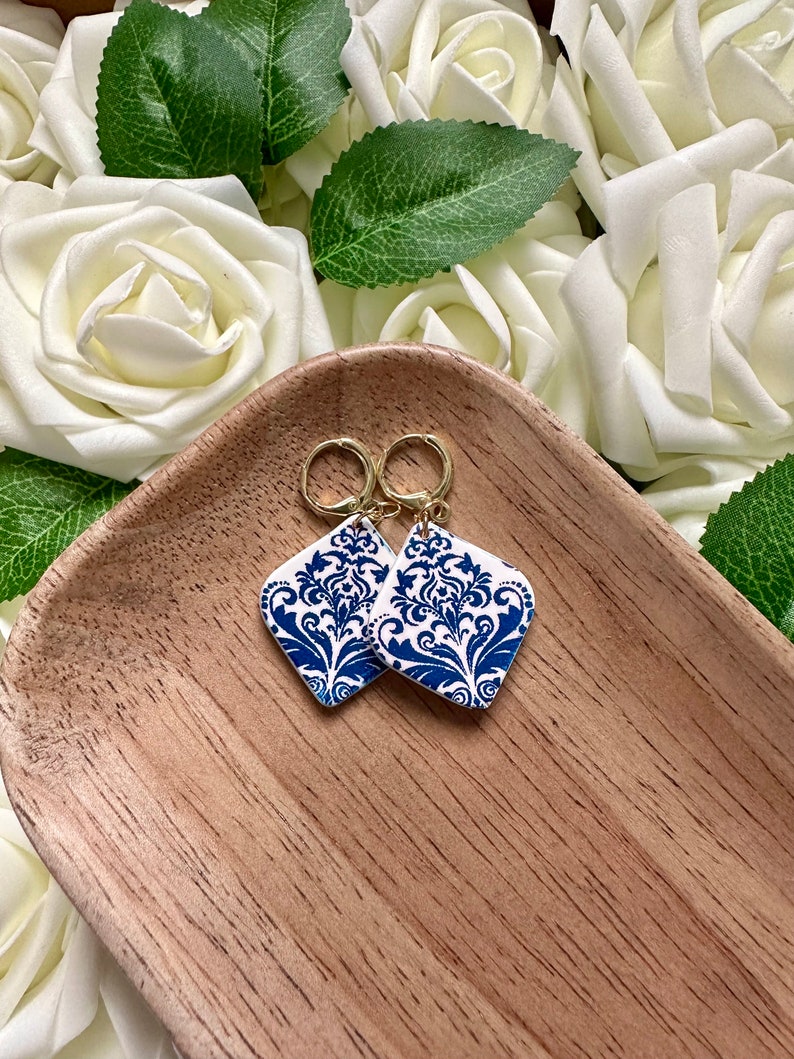 May include: A pair of blue and white patterned earrings with gold hoops. The earrings are shaped like a diamond and feature a floral design.