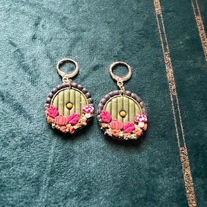 May include: A pair of green and brown earrings featuring a round, green door design. The door has a small, round, gold knob and is surrounded by small, orange pumpkins, red mushrooms, and brown leaves.