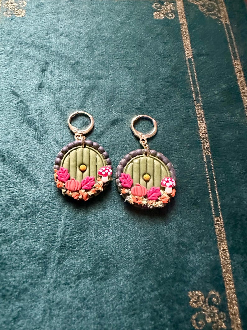 May include: A pair of green and brown earrings featuring a round, green door design. The door has a small, round, gold knob and is surrounded by small, orange pumpkins, red mushrooms, and brown leaves.