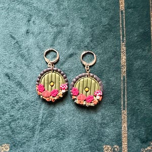 May include: A pair of green and brown earrings with a round door design, featuring a small yellow circle in the centre and a wreath of leaves, mushrooms, and pumpkins around the door.