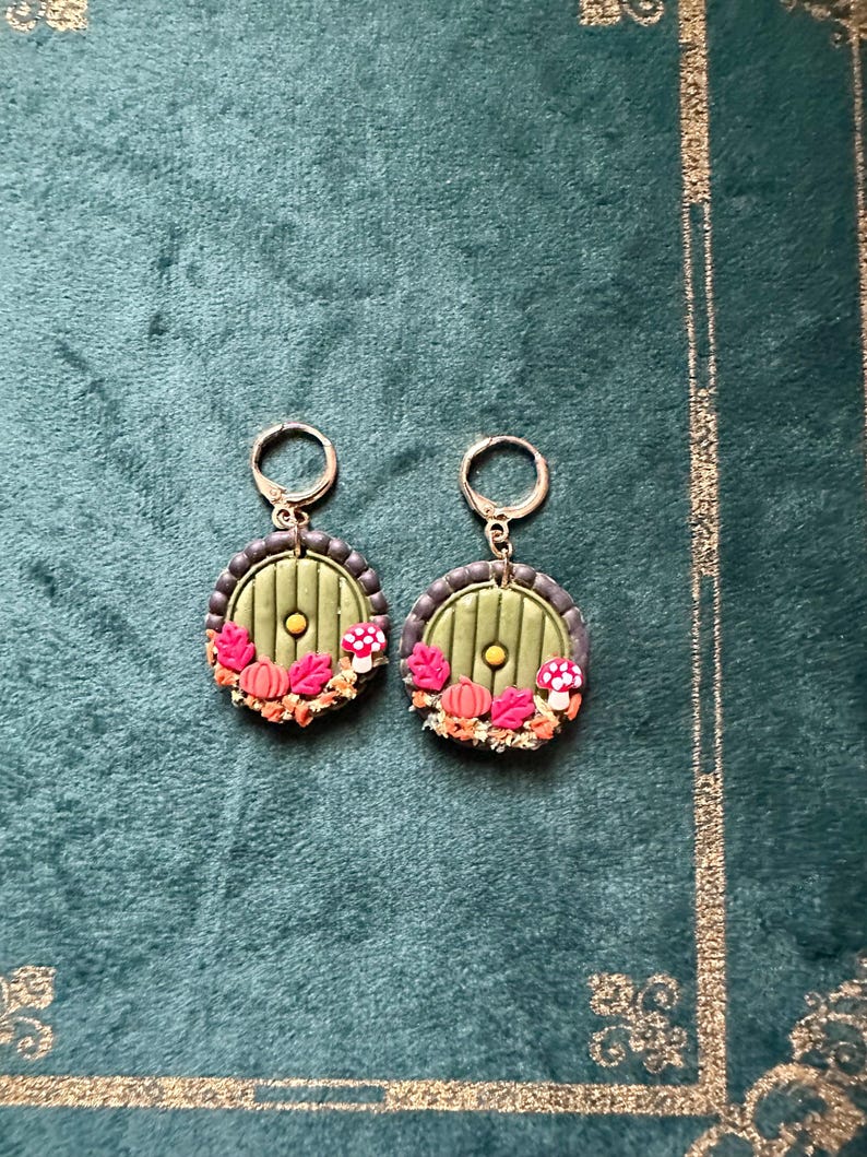 May include: A pair of green and brown earrings with a round door design, featuring a small yellow circle in the centre and a wreath of leaves, mushrooms, and pumpkins around the door.