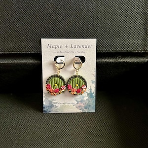 May include: A pair of green and gold earrings featuring a miniature replica of the hobbit hole door from The Lord of the Rings. The door is surrounded by autumn leaves, pumpkins, and mushrooms.