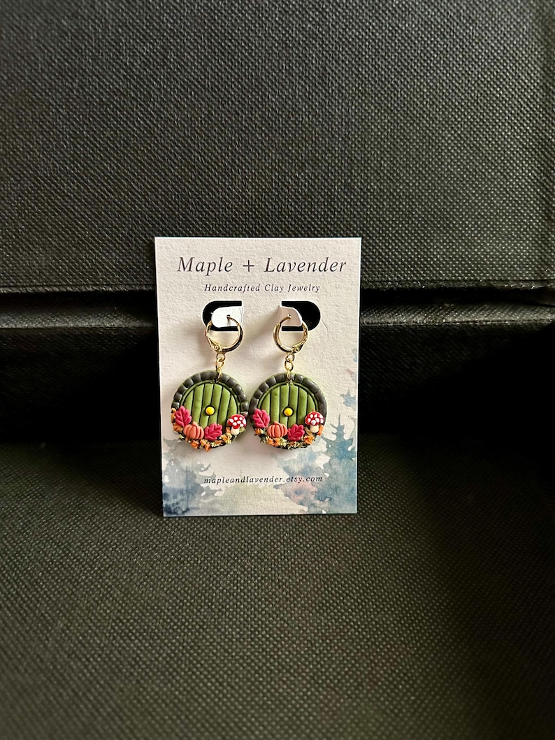 May include: A pair of green and gold earrings featuring a miniature replica of the hobbit hole door from The Lord of the Rings. The door is surrounded by autumn leaves, pumpkins, and mushrooms.
