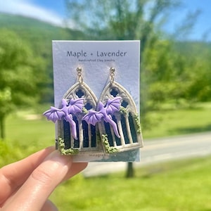 May include: Handcrafted clay jewelry earrings featuring a gothic window design with purple dragon accents. The earrings are detailed with green moss and gold-colored hooks. The earrings are displayed on a card with the text "Maple + Lavender" and "Instagram".