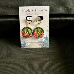 May include: A pair of green and gold earrings featuring a round, green door design. The door has a small, yellow window and is surrounded by red and orange flowers. The earrings are handcrafted and are available for purchase on Etsy.