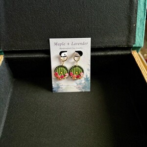 May include: A pair of green and gold earrings with a round, door-shaped design. The earrings are decorated with pink and red flowers. The earrings are displayed on a white card with the text "Maple + Lavender Handmade Clay Jewelry".