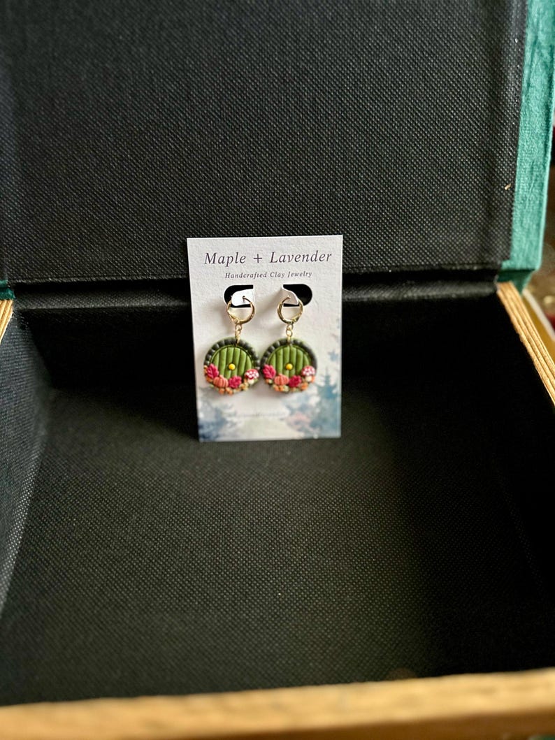 May include: A pair of green and gold earrings with a round, door-shaped design. The earrings are decorated with pink and red flowers. The earrings are displayed on a white card with the text "Maple + Lavender Handmade Clay Jewelry".