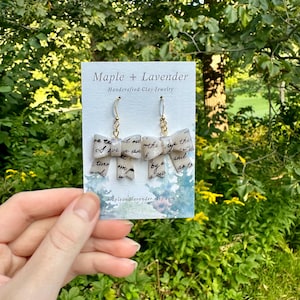 May include: A pair of white clay earrings shaped like bows with handwritten text on them. The earrings are on a white card with the text "Maple + Lavender Handcrafted Clay Jewelry" and a website address.