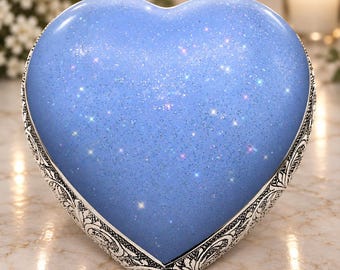 Puede incluir: Una urna en forma de corazón con un acabado azul claro liso y un efecto brillante que imita un cielo estrellado. Los bordes del corazón están enmarcados por un borde decorativo de metal plateado con un intrincado patrón floral en relieve.