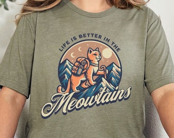 May include: A green t-shirt with a cat wearing a backpack and hiking in the mountains. The text on the shirt reads "Life is better in the Meowntains".