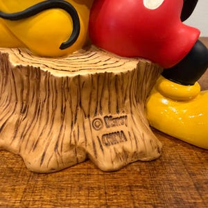 K&ouml;nnte beinhalten: Eine Keramikfigur mit Micky Maus und Pluto. Mickys rote Shorts und schwarzen Schuhe sind zu sehen, zusammen mit Plutos gelbem K&ouml;rper. Die Basis ist ein Baumstumpf mit den Disney- und China-Marken.