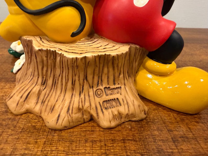 K&ouml;nnte beinhalten: Eine Keramikfigur mit Micky Maus und Pluto. Mickys rote Shorts und schwarzen Schuhe sind zu sehen, zusammen mit Plutos gelbem K&ouml;rper. Die Basis ist ein Baumstumpf mit den Disney- und China-Marken.
