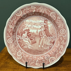 Peut inclure: Une assiette décorative avec un bord festonné et une bordure florale détaillée. Le centre présente une scène pastorale sépia avec des personnages et une église. L'assiette est soutenue par un support en métal noir, mettant en valeur son design.