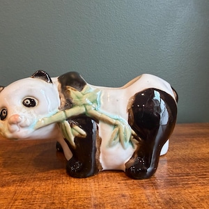 Puede incluir: Jardinera de cerámica con forma de oso panda. El panda tiene un cuerpo blanco, orejas negras y patas negras. Presenta una cara sonriente y una rama de bambú verde. La jardinera está sobre una superficie de madera.