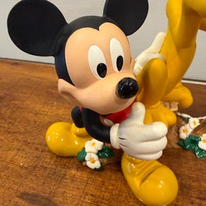 K&ouml;nnte beinhalten: Eine Mickey Mouse-Figur mit schwarzen Ohren, einem beigen Gesicht und einem roten Hemd. Mickey tr&auml;gt gelbe Schuhe und wei&szlig;e Handschuhe und zeigt den Daumen hoch. Die Figur ist mit kleinen wei&szlig;en Blumen verziert.