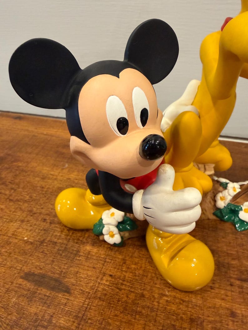 K&ouml;nnte beinhalten: Eine Mickey Mouse-Figur mit schwarzen Ohren, einem beigen Gesicht und einem roten Hemd. Mickey tr&auml;gt gelbe Schuhe und wei&szlig;e Handschuhe und zeigt den Daumen hoch. Die Figur ist mit kleinen wei&szlig;en Blumen verziert.