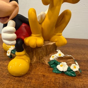K&ouml;nnte beinhalten: Eine farbenfrohe Figur mit Micky Maus und Pluto. Micky tr&auml;gt seine klassische Kleidung mit roter Hose, schwarzen Armen und gelben Schuhen. Pluto ist gelb. Der Sockel ist ein Baumstumpf mit Blumen.