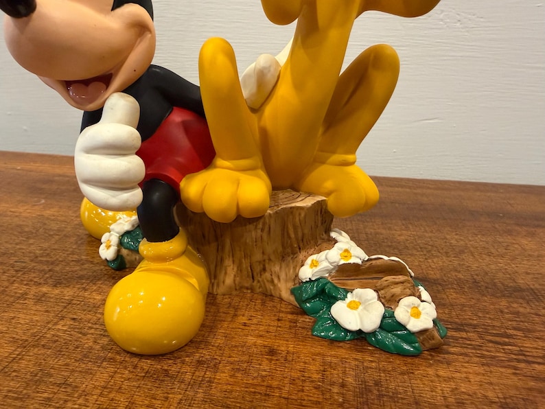 K&ouml;nnte beinhalten: Eine farbenfrohe Figur mit Micky Maus und Pluto. Micky tr&auml;gt seine klassische Kleidung mit roter Hose, schwarzen Armen und gelben Schuhen. Pluto ist gelb. Der Sockel ist ein Baumstumpf mit Blumen.