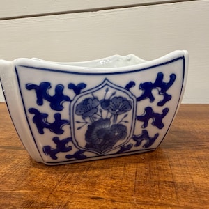 Peut inclure: Un bol en porcelaine blanc et bleu avec un motif floral. Le bol a une forme rectangulaire avec des côtés incurvés et un motif floral bleu sur le devant. Le bol est posé sur une surface en bois.