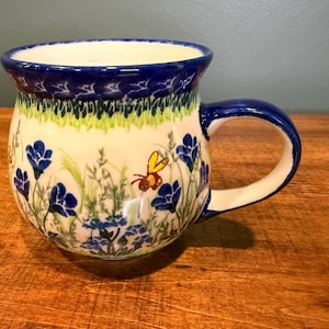 May include: A ceramic mug with a blue rim and handle, decorated with a floral and bee design. The mug features blue flowers, green grass, and a yellow and brown bee. The background is cream-coloured.