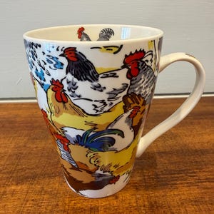 May include: White ceramic mug with a wrap-around design featuring colorful illustrations of chickens and roosters. The mug has a curved handle and a wide opening. The rooster illustrations include red, black, white, and yellow colors.