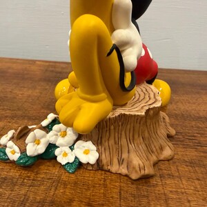 K&ouml;nnte beinhalten: Eine Keramikfigur mit Micky Maus und Pluto. Micky tr&auml;gt wei&szlig;e Handschuhe, rote Shorts und schwarze Schuhe. Pluto hat gelbe Beine und F&uuml;&szlig;e. Die Figur steht auf einem Baumstumpf mit Blumen.